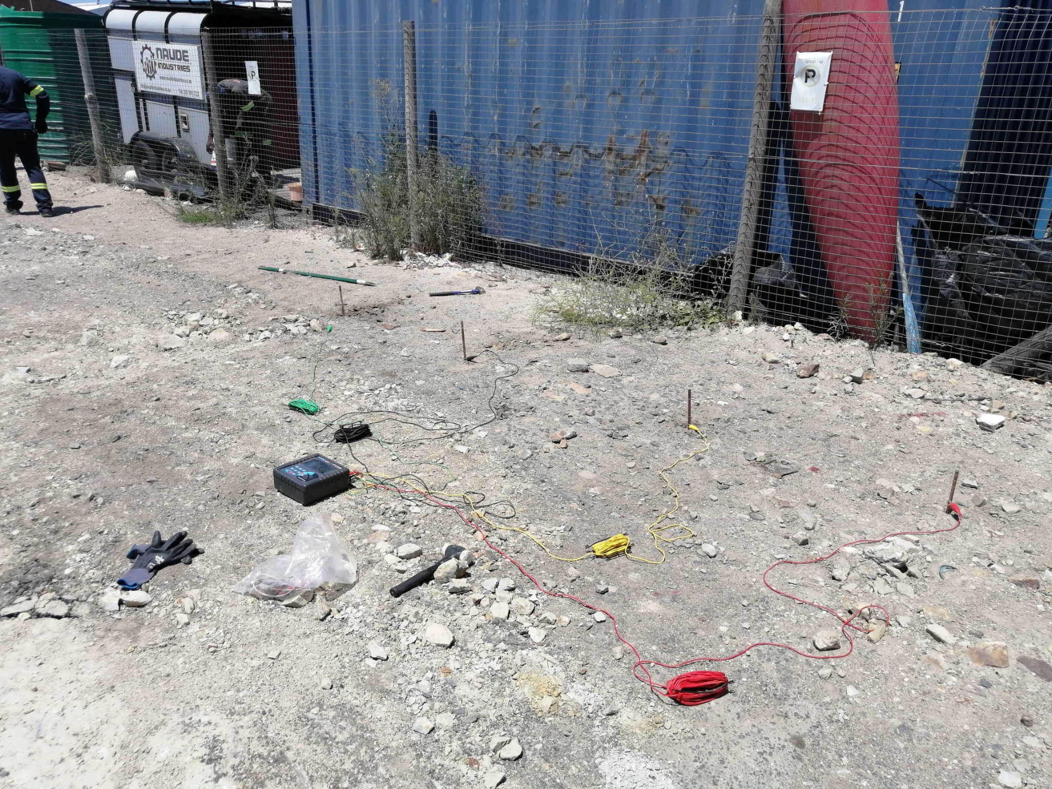 Test stakes and colour-coded leads laid out on gravel beside an industrial fence